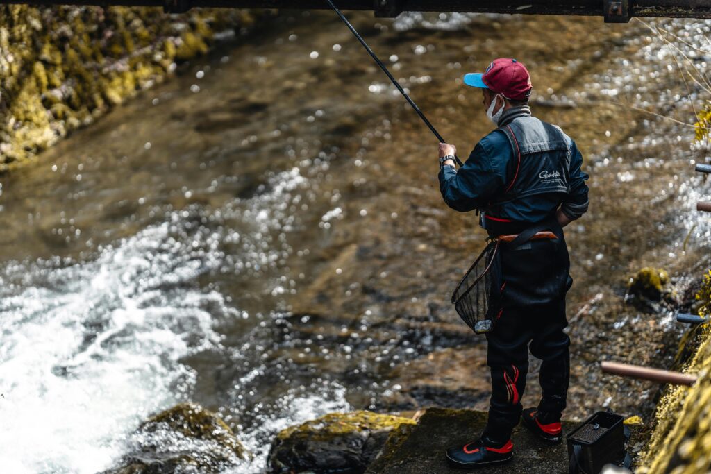 A person in fishing gear casting a fishing line by a river on a sunny day.